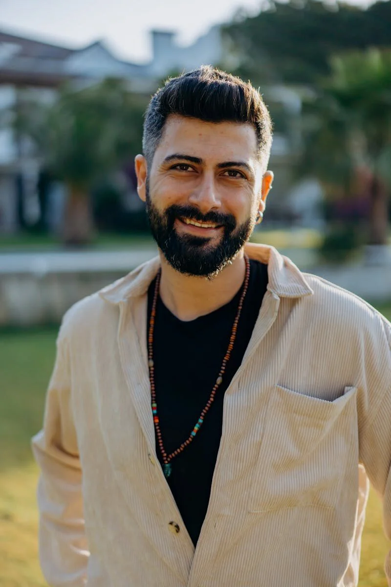 A cheerful man with a beard smiling outdoors in a sunlit garden setting.