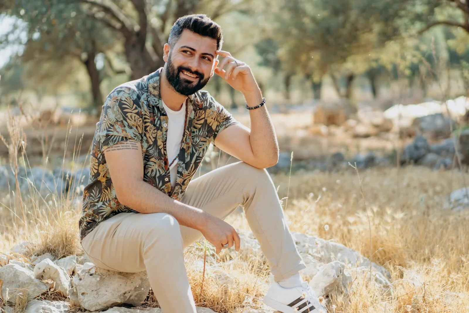 A fashionable man with a beard smiling while sitting outdoors in sunny nature.