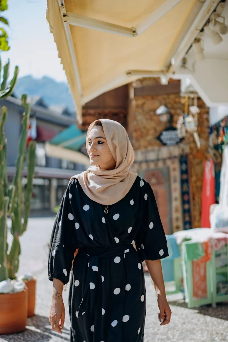 Smiling woman wearing hijab stands in a sunny outdoor market surrounded by vibrant textiles and greenery.